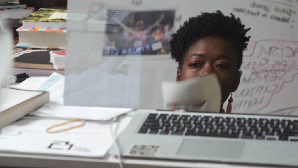 Racismo algorítmico: pesquisadora Joy Buolamwini está diante de um computador segurando uma máscara branca que cobre metade de seu rosto.